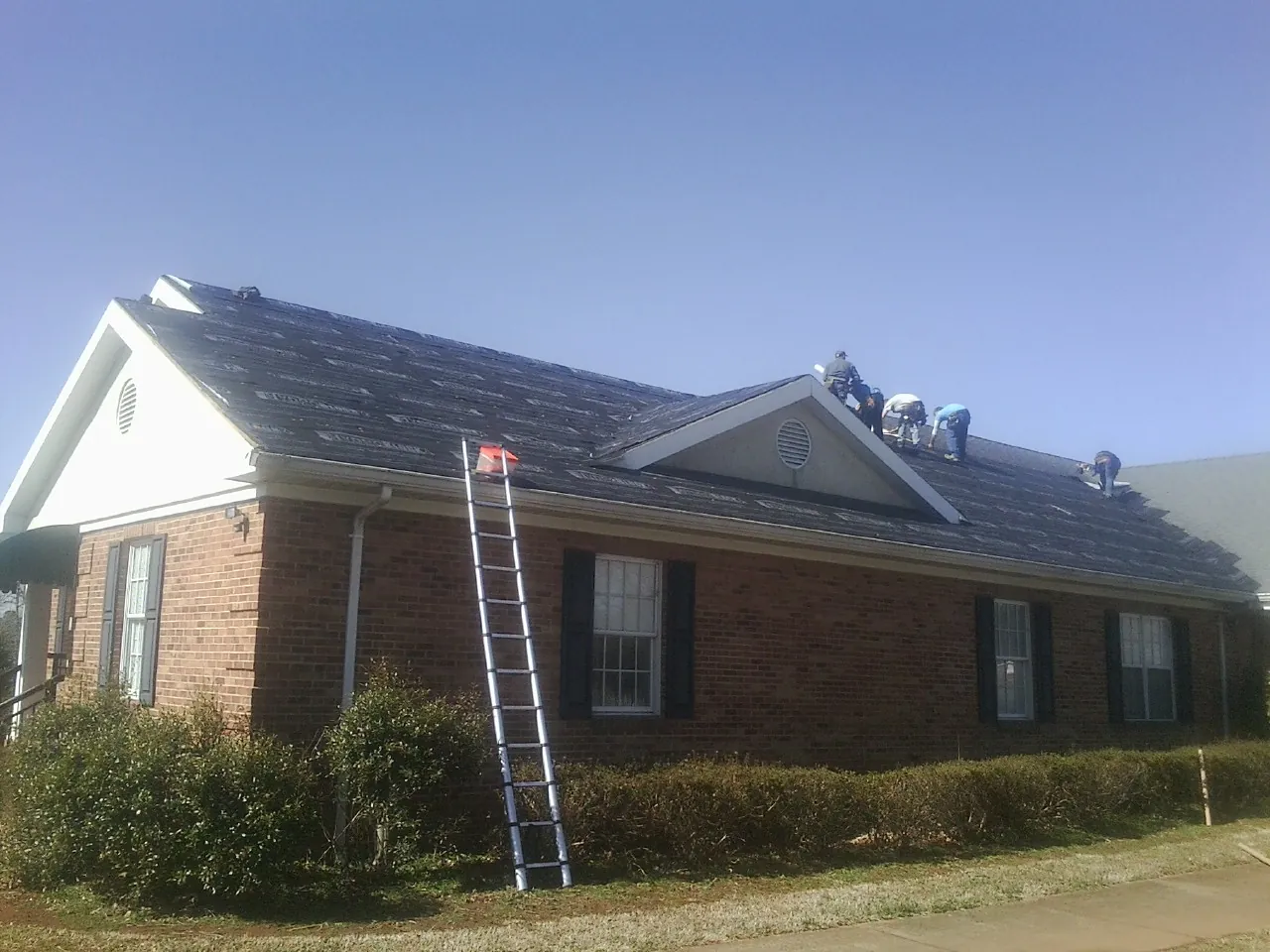 Crew tearing shingles off a brick ranch roof with a ladder set — Metal Roof Repair work by our crew in New Iberia