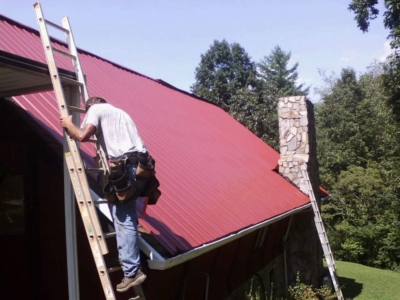 Roofer on a ladder beside a red metal roof and stone chimney — Soffit & Fascia Repair by Roofing Brothers in New Iberia